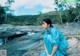 A woman in a blue dress sitting on a rock by a waterfall.